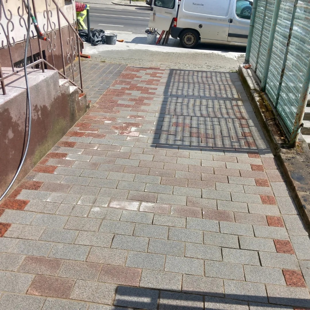 Świeżo ułożony chodnik z szarej kostki z czerwonymi akcentami, częściowo mokry po czyszczeniu, widoczny pracownik i samochód dostawczy w tle.