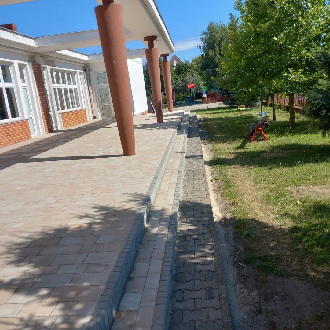 Jasna kostka brukowa ułożona na zewnątrz budynku z kolumnami, obok widać obrzeże z szarej kostki brukowej i fragment placu zabaw.