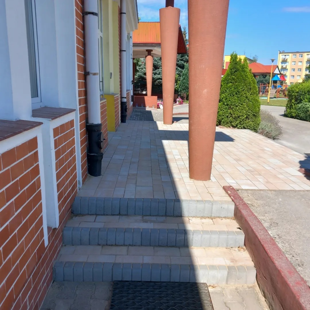Chodnik z jasnoszarej kostki brukowej prowadzący wzdłuż budynku z ceglaną elewacją i kolumnami w kolorze terakoty, widoczne schody z kostki i elementy infrastruktury miejskiej w tle.