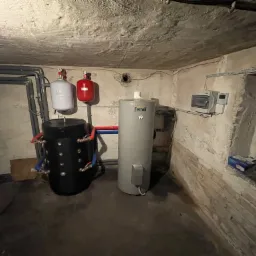 Zestaw instalacji grzewczej w piwnicy: czarny zbiornik buforowy z licznymi przyłączami, szary piec Ferroli, biały i czerwony zbiornik wyrównawczy, orurowanie pod sufitem, regulator temperatury...