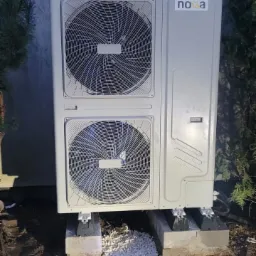 Zewnętrzna jednostka pompy ciepła marki Noxa, umieszczona na betonowych podstawach z podsypką z białego żwiru, na tle szarej ściany i zieleni.