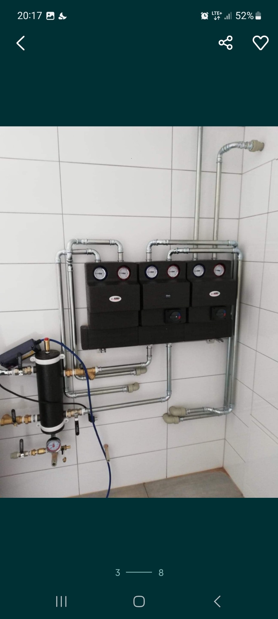 Złożony system instalacji grzewczej z czarnymi modułami, manometrami i licznymi rurami na tle białych płytek.