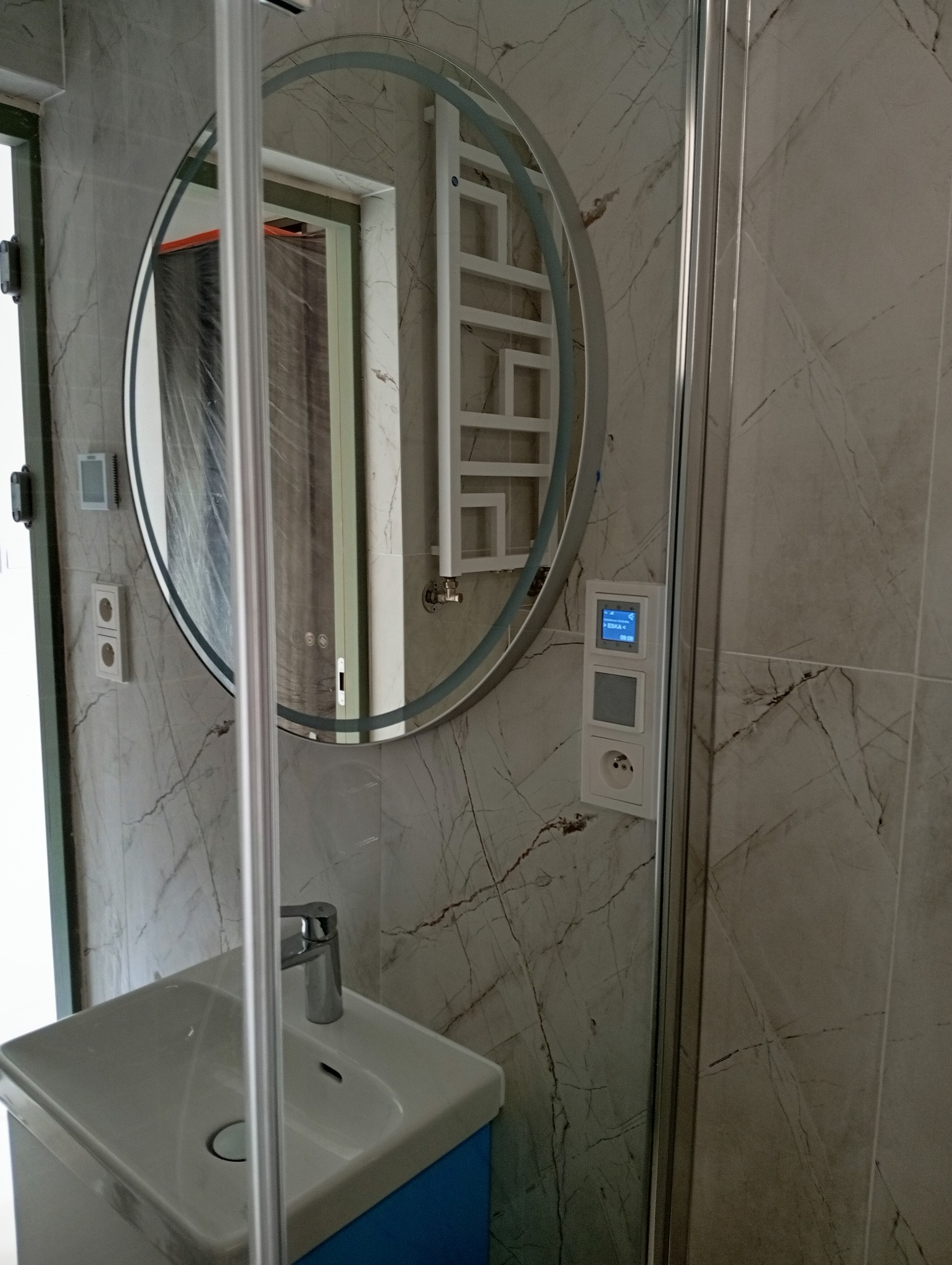 Nowoczesna łazienka z umywalką na niebieskiej szafce, okrągłym lustrem z podświetleniem LED, grzejnikiem drabinkowym i marmurowymi płytkami.