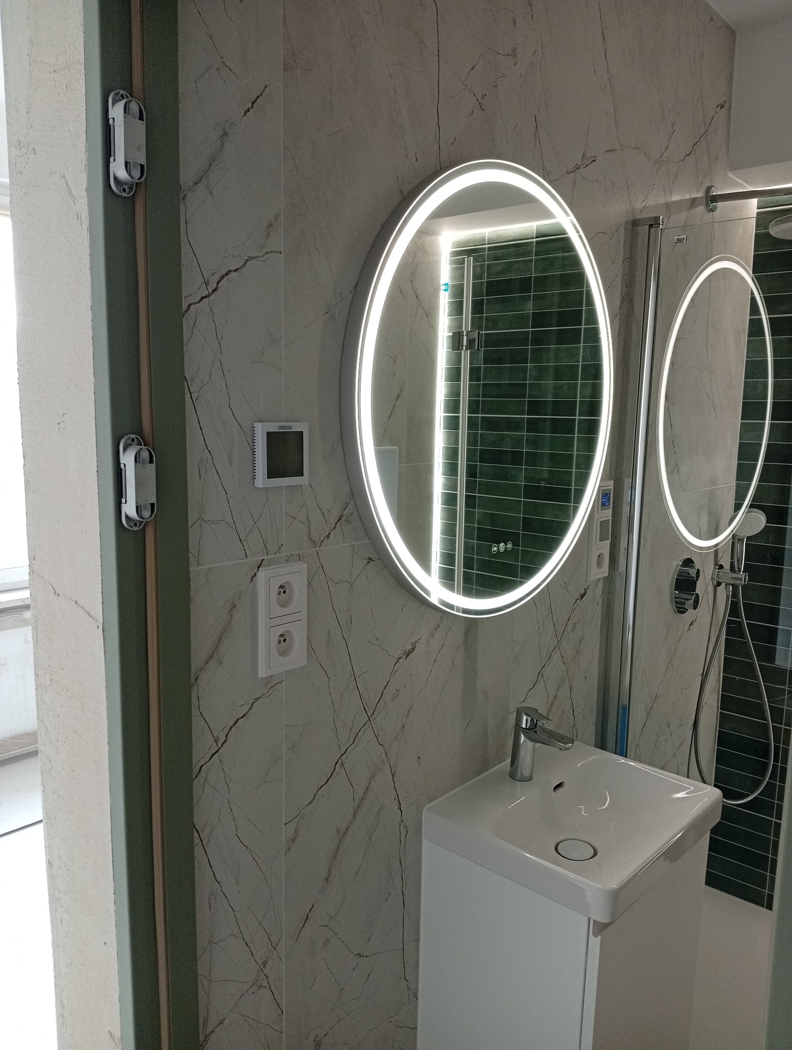 Nowoczesna łazienka z okrągłym lustrem LED, umywalką na szafce, marmurowymi płytkami ściennymi i kabiną prysznicową z zielonymi płytkami w tle.