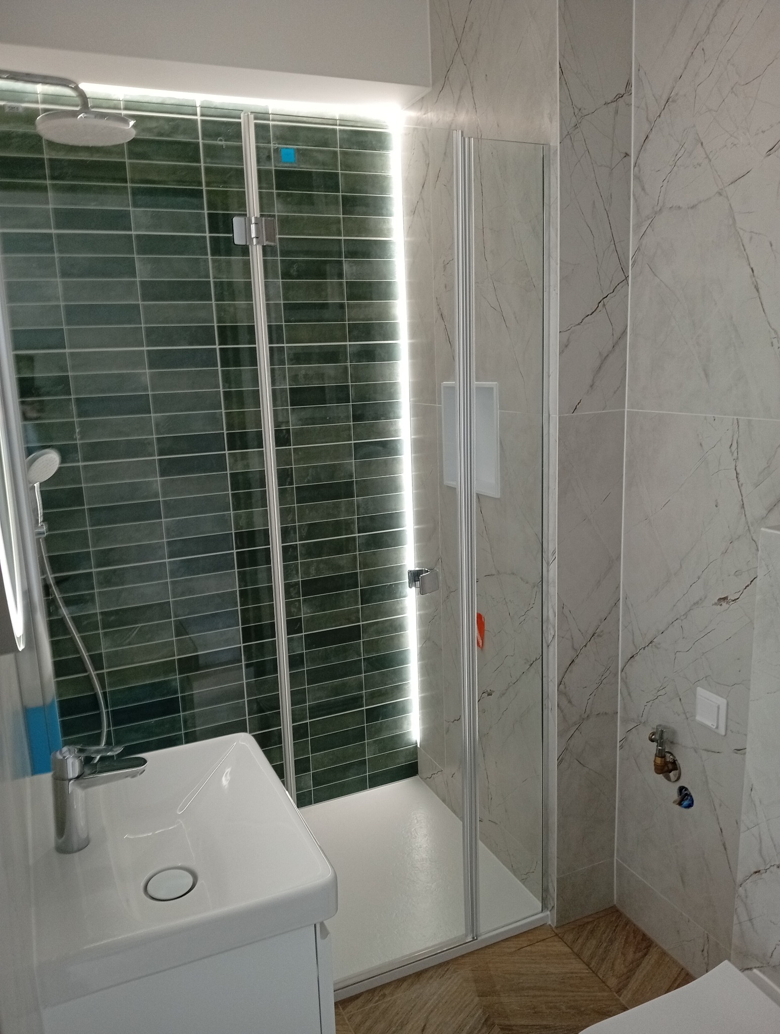 Nowoczesna łazienka z kabiną prysznicową ze szklanymi drzwiami, umywalką na szafce, zielonymi płytkami w strefie prysznica i marmurowymi płytkami na ścianie obok.