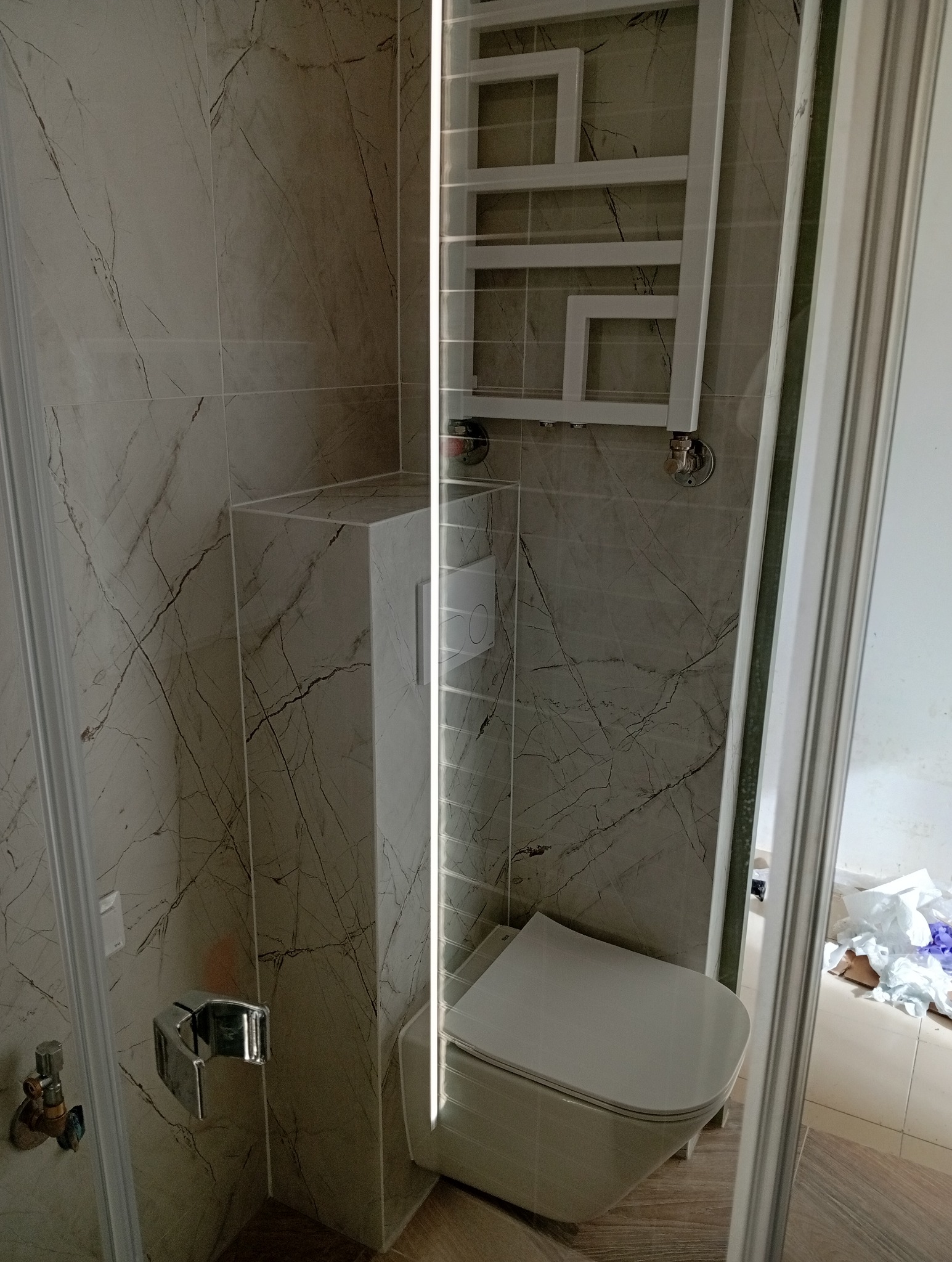 Nowoczesna toaleta podwieszana z szarymi płytkami imitującymi marmur, chromowany uchwyt na papier toaletowy i biały grzejnik drabinkowy w łazience po remoncie.