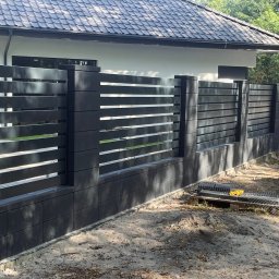Superpłot - Nowoczesne, poziome ogrodzenie metalowe w kolorze antracytowym, z elementami betonowymi, otaczające posesję z domem o ciemnej dachówce. Widok z poziomu gruntu.