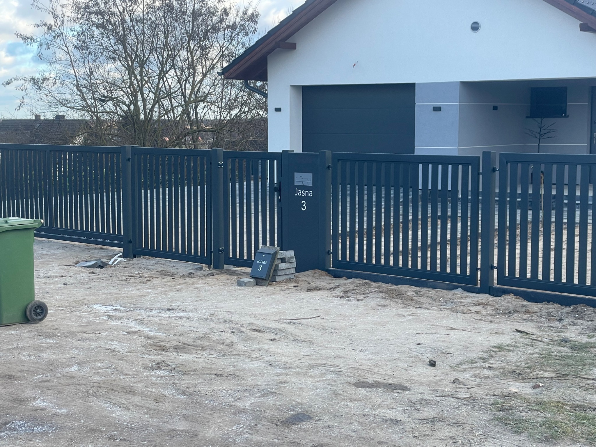 Nowoczesne, grafitowe ogrodzenie panelowe z furtką i numerem domu 'Jasna 3' na tle minimalistycznego budynku. Widoczne niedokończone prace brukarskie.