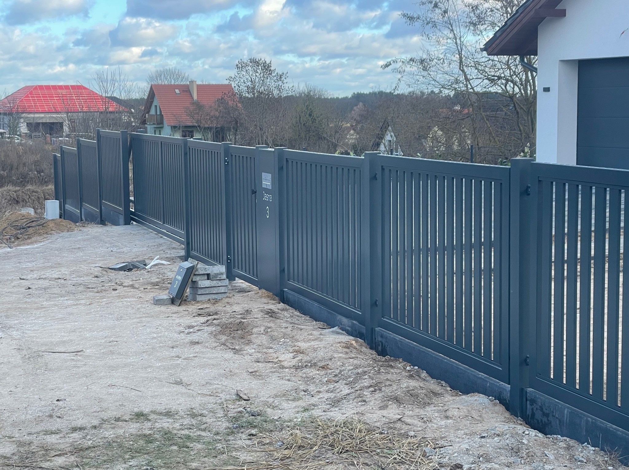 Nowoczesna, grafitowa brama przesuwna z pionowymi szczebelkami, zamontowana przy posesji z domem i budynkiem w trakcie budowy w tle. Widoczne bloczki betonowe.