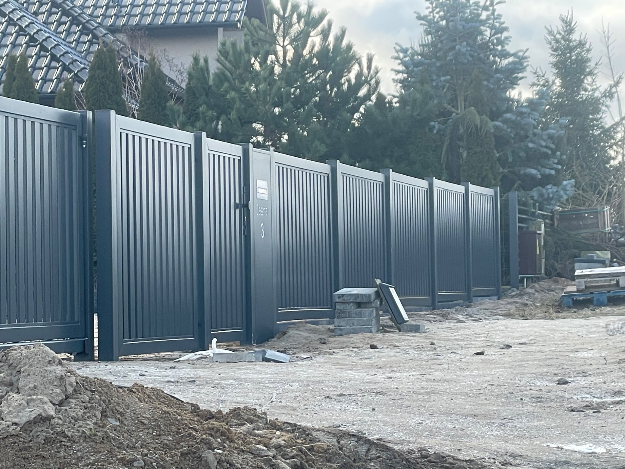 Nowoczesne, szare ogrodzenie panelowe z bramą wjazdową na tle zieleni. Widoczne elementy montażowe i teren po budowie. Ujęcie z bliska, podkreślające detale.