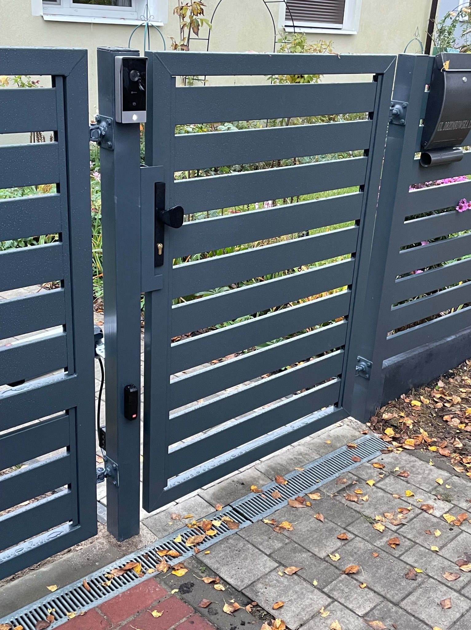 Grafitowa brama wjazdowa z poziomymi panelami, wyposażona w wideodomofon i skrzynkę na listy, zamontowana na posesji z kostką brukową i odpływem liniowym.