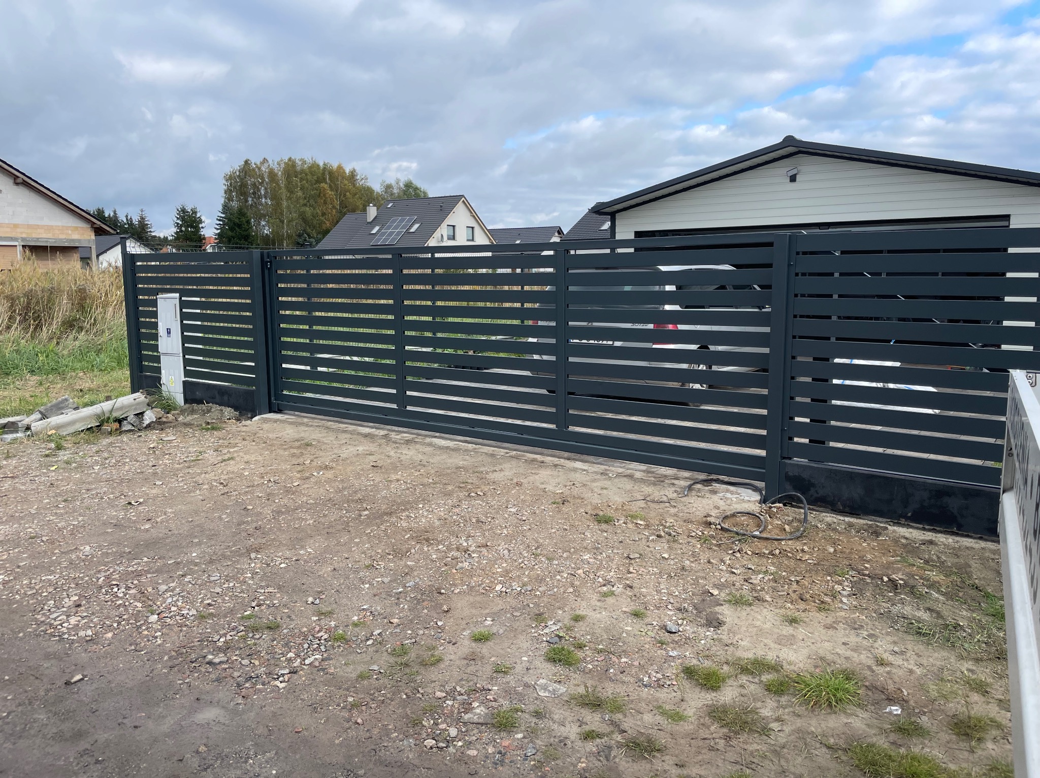 Nowoczesna, przesuwna brama wjazdowa z poziomych, grafitowych paneli na tle domu jednorodzinnego i pochmurnego nieba. Widoczne elementy instalacji elektrycznej.