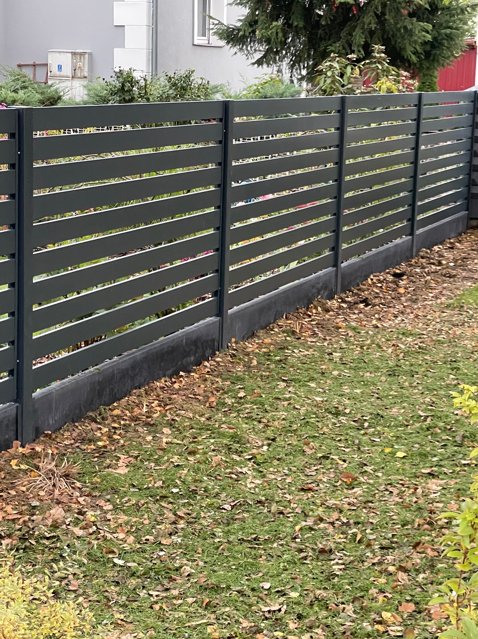 Nowoczesne, poziome ogrodzenie metalowe w kolorze grafitowym, oddzielające posesję od zielonego trawnika z opadłymi liśćmi. Widok na dom w tle.