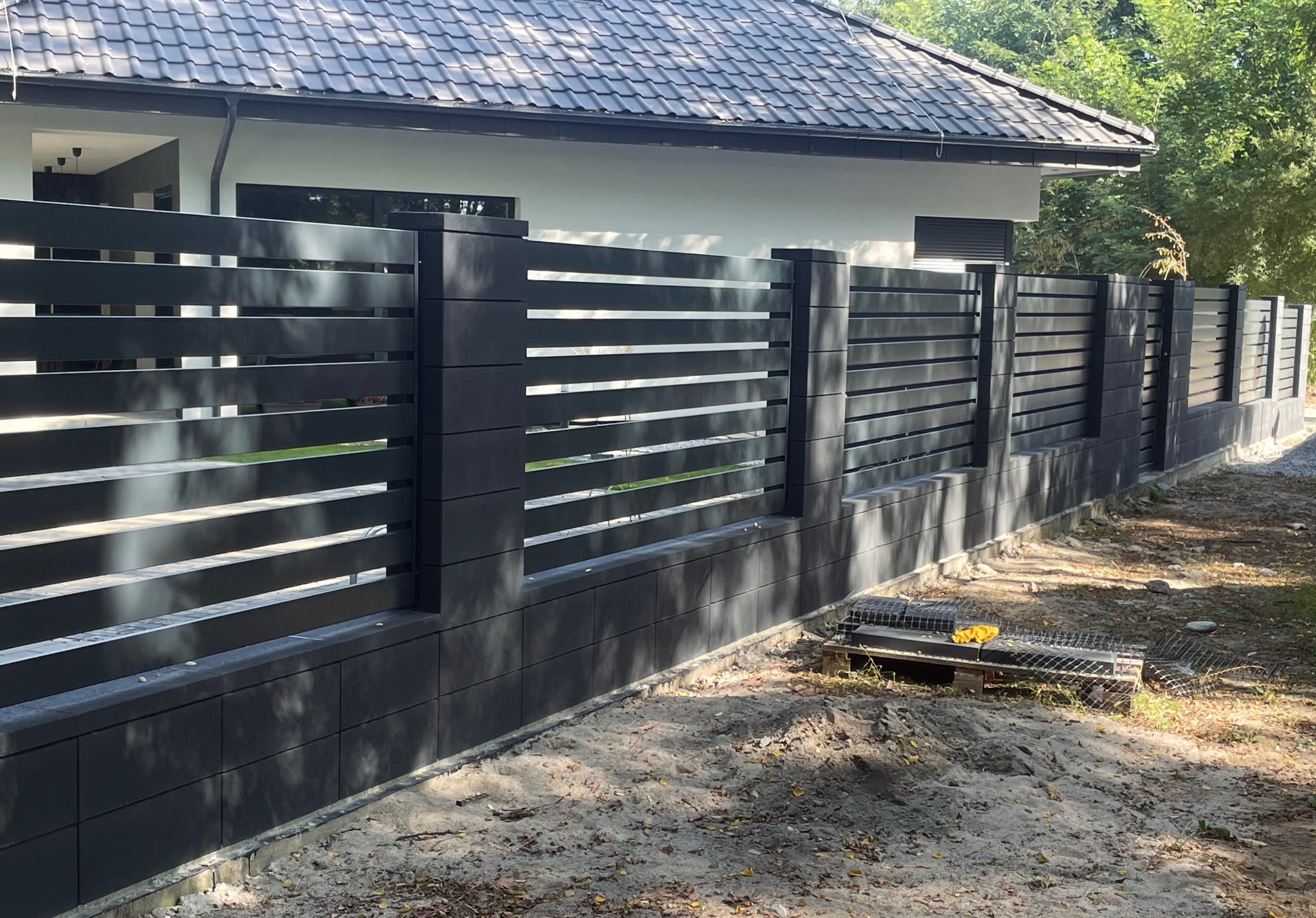 Nowoczesne, poziome ogrodzenie metalowe w kolorze antracytowym, z elementami betonowymi, otaczające posesję z domem jednorodzinnym. Widok z zewnątrz.