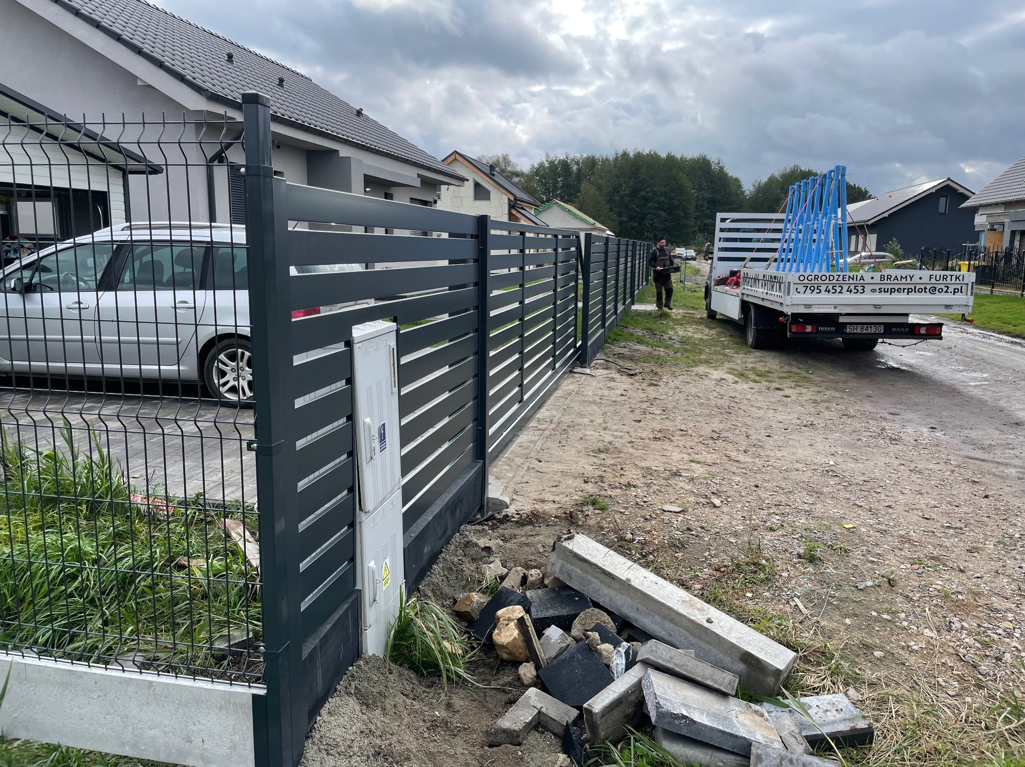 Nowoczesne, grafitowe ogrodzenie panelowe z bramą przesuwną, widok z zewnątrz posesji, z dostawczym autem w tle. Częściowo ukończony montaż.