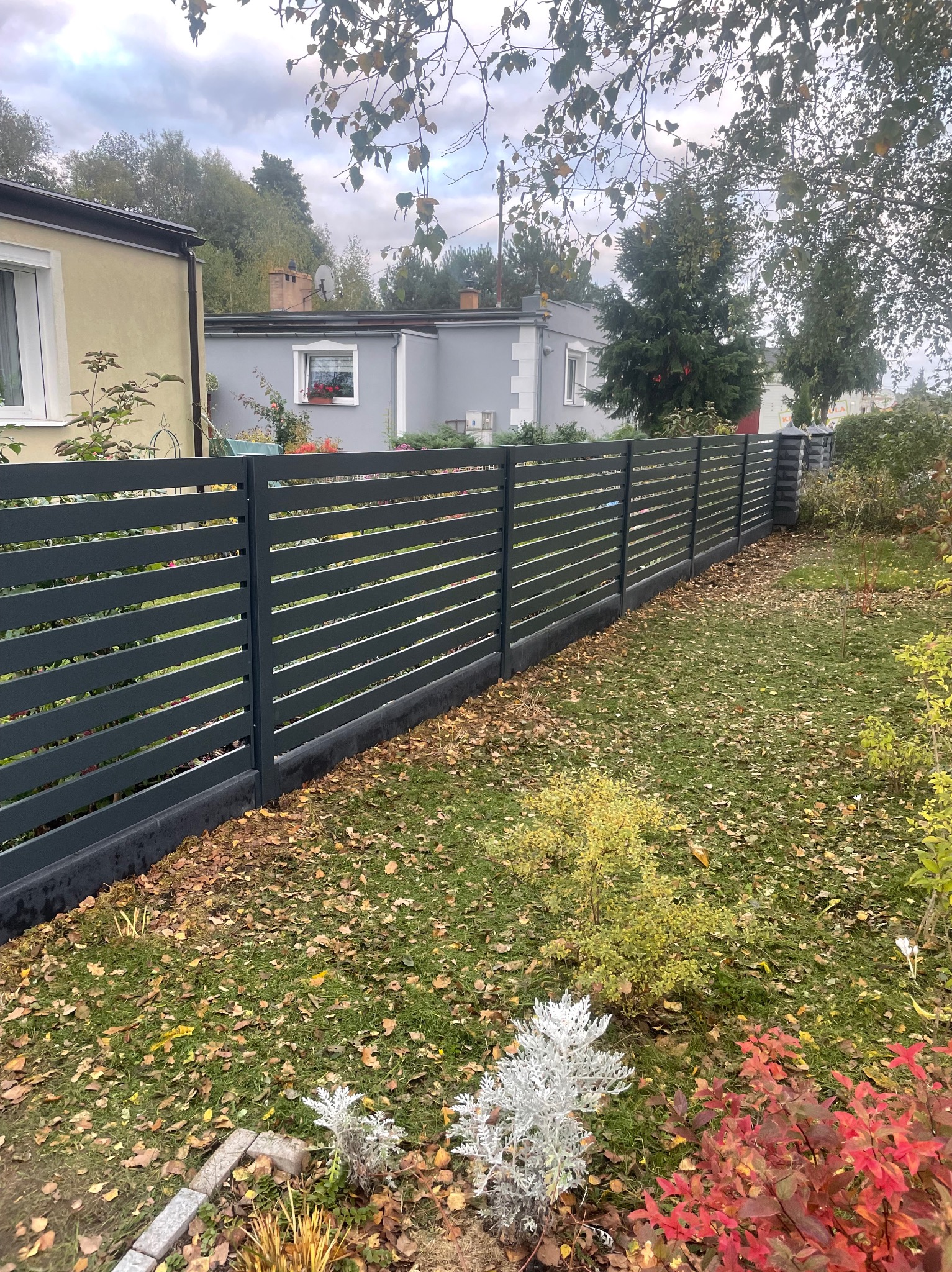 Nowoczesne, poziome ogrodzenie panelowe w kolorze grafitowym, oddzielające posesję z domem w tle. Jesienne liście na trawie dodają koloru.