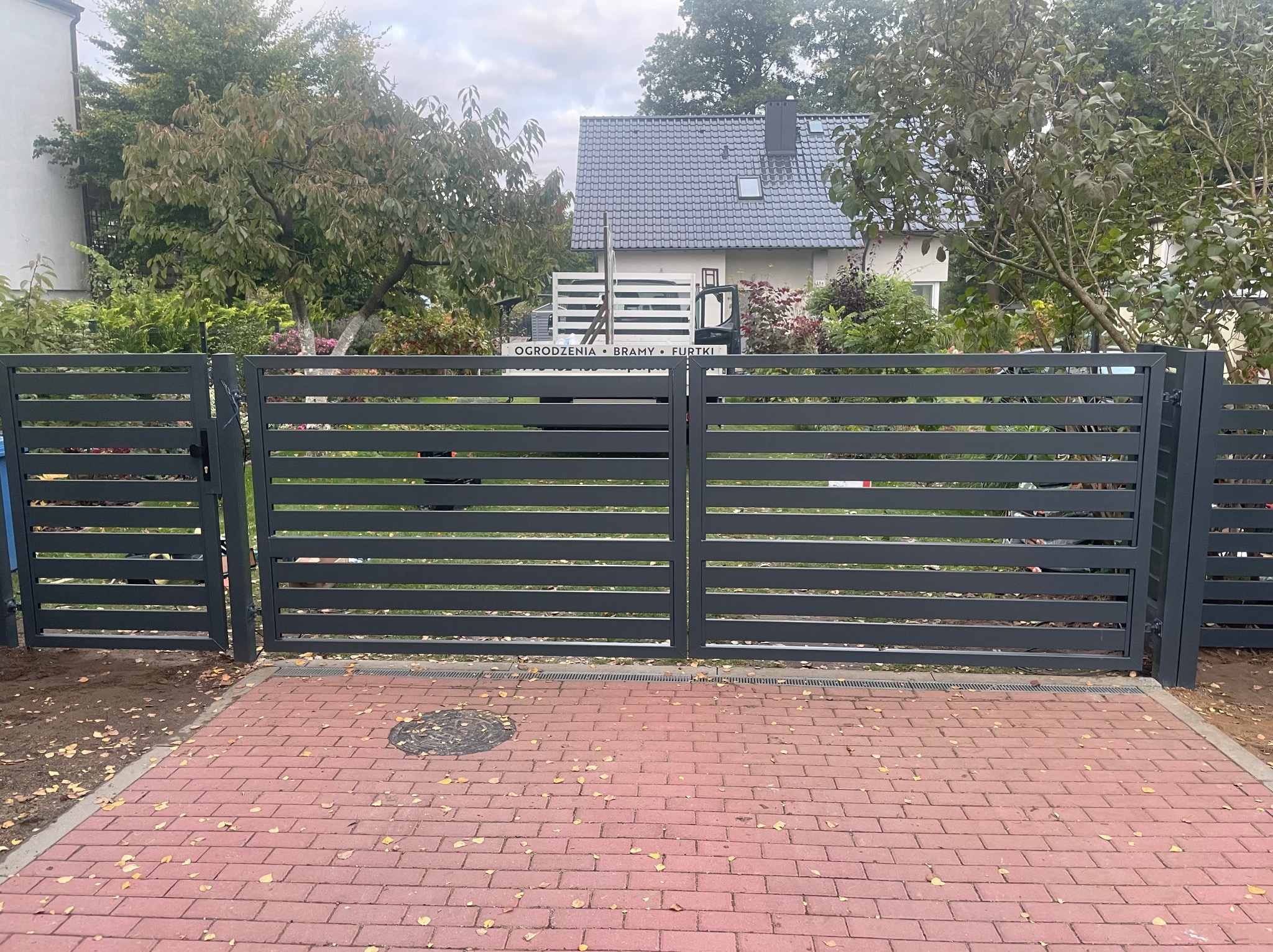 Nowoczesna, pozioma brama metalowa w kolorze grafitowym, z furtką po lewej stronie. Czerwona kostka brukowa na podjeździe, dom z szarą dachówką w tle.