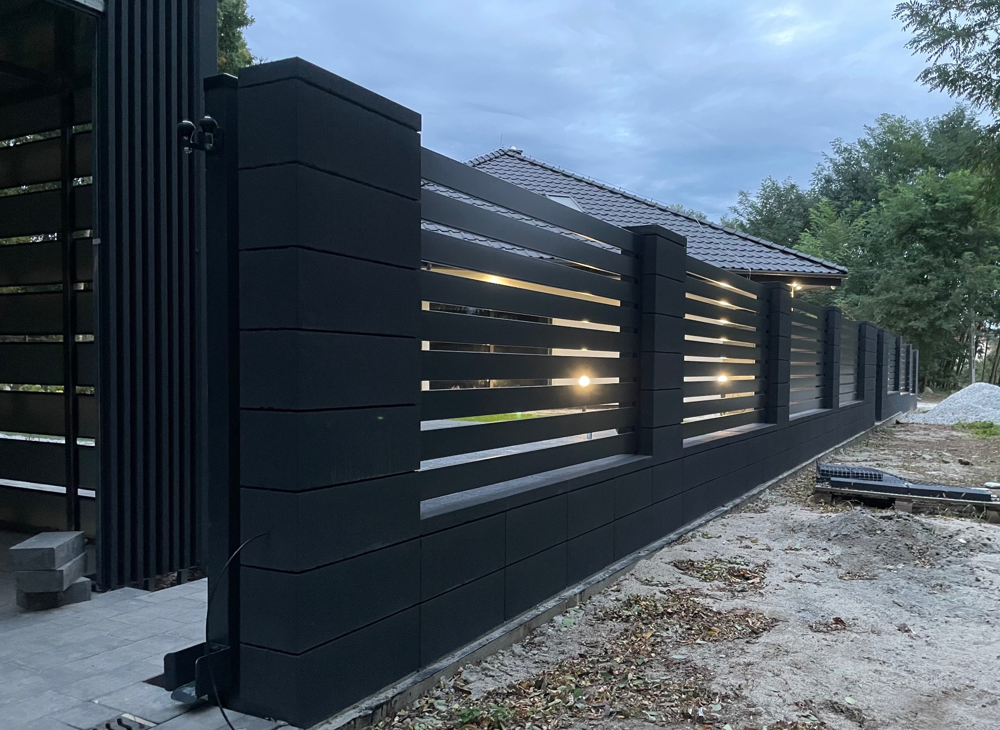 Nowoczesne, czarne ogrodzenie metalowe z poziomymi panelami i wbudowanym oświetleniem, widoczne z boku, z elementami automatyki do bram w tle.