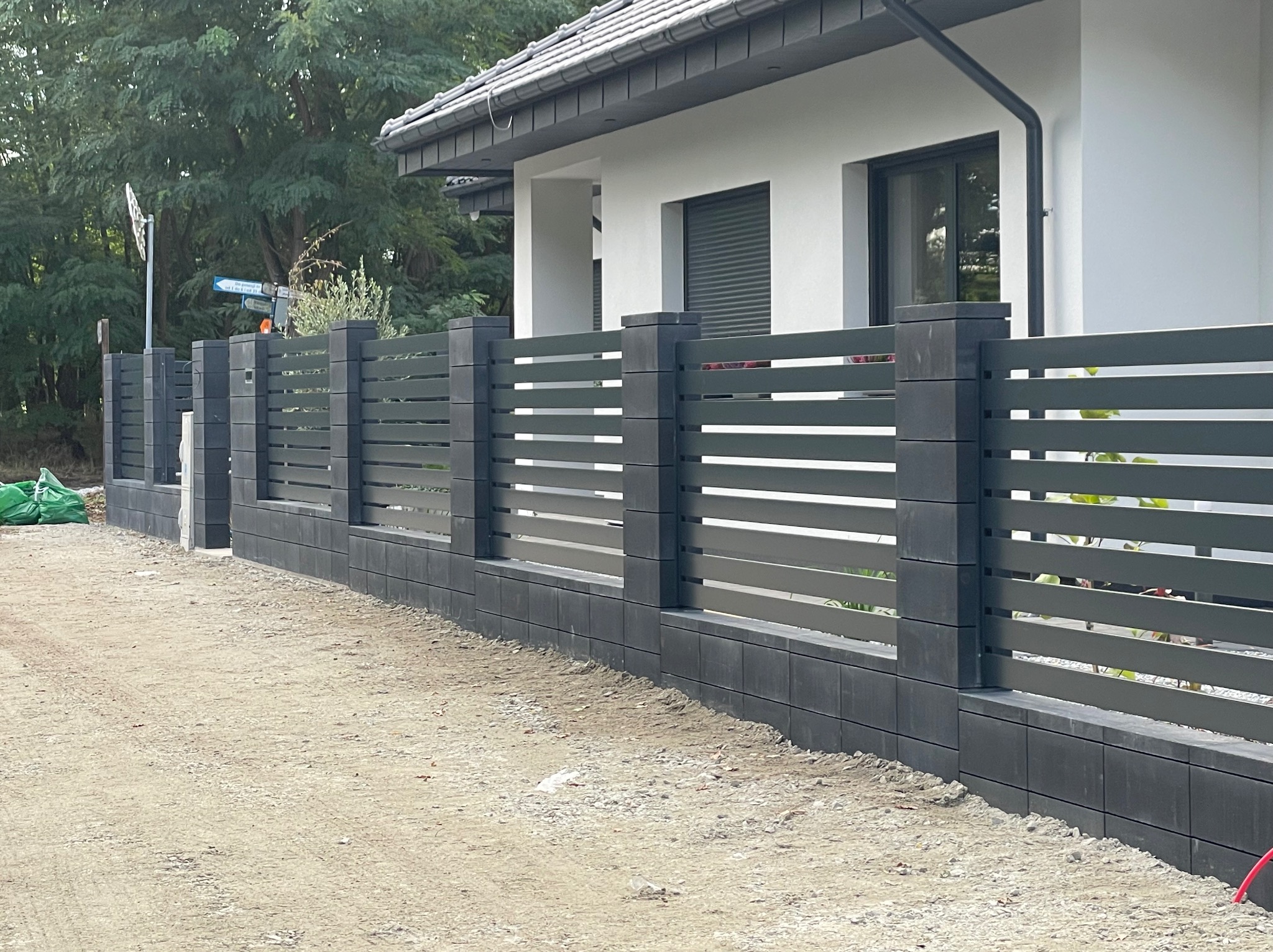 Nowoczesne ogrodzenie panelowe z poziomymi przęsłami w kolorze grafitowym, na niskiej podmurówce z bloczków, na tle białego domu z szarą dachówką.