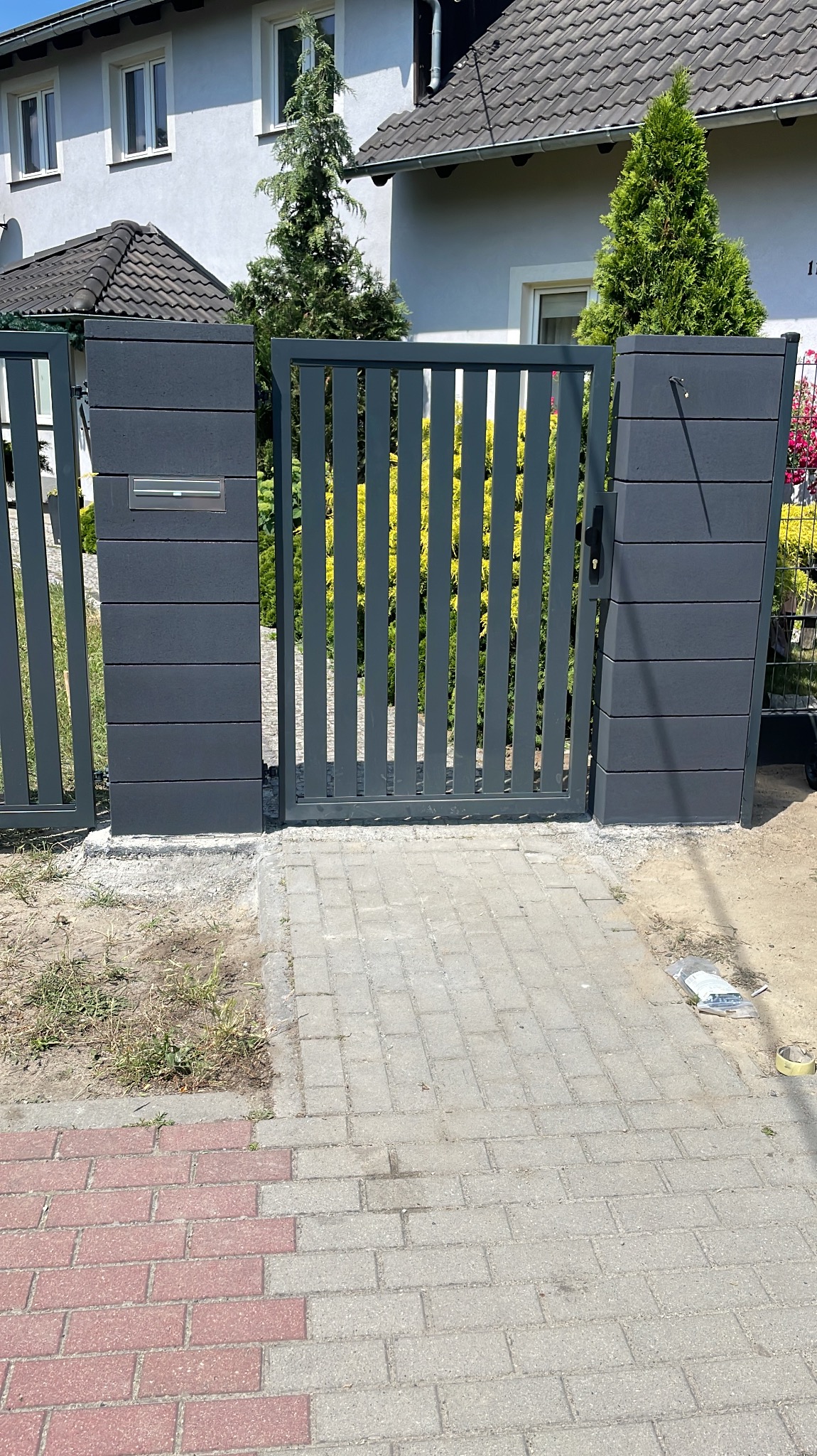 Nowoczesna, grafitowa furtka z pionowych sztachet, wbudowana w ogrodzenie z paneli kompozytowych. Widoczna skrzynka na listy i kostka brukowa.