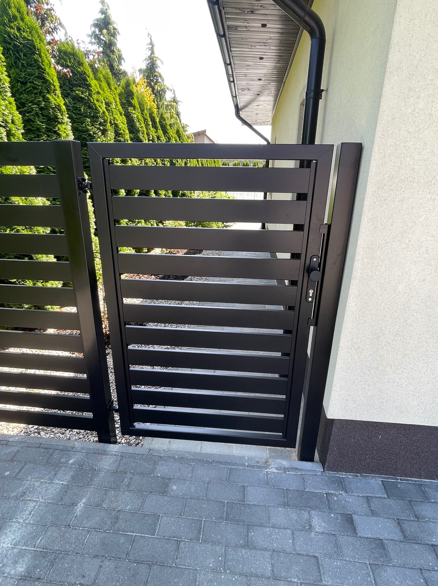 Nowoczesna, czarna brama metalowa z poziomymi szczeblami, widoczna z bliska na tle zieleni i kostki brukowej. Zamek z czarną gałką po prawej stronie.