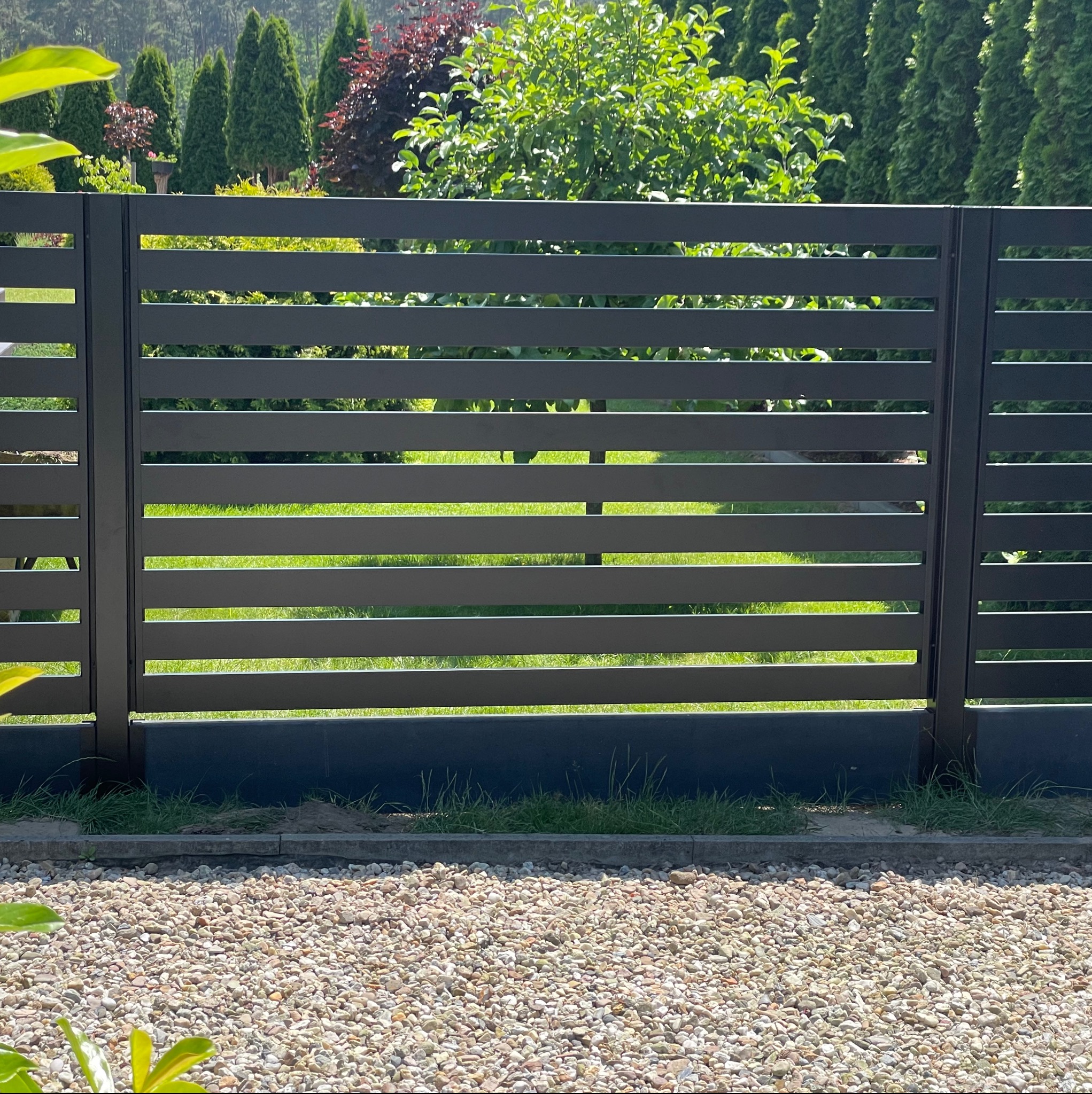 Nowoczesne, poziome ogrodzenie panelowe w kolorze grafitowym, z widokiem na zielony trawnik i drzewa w tle. Minimalistyczny design i staranne wykonanie.