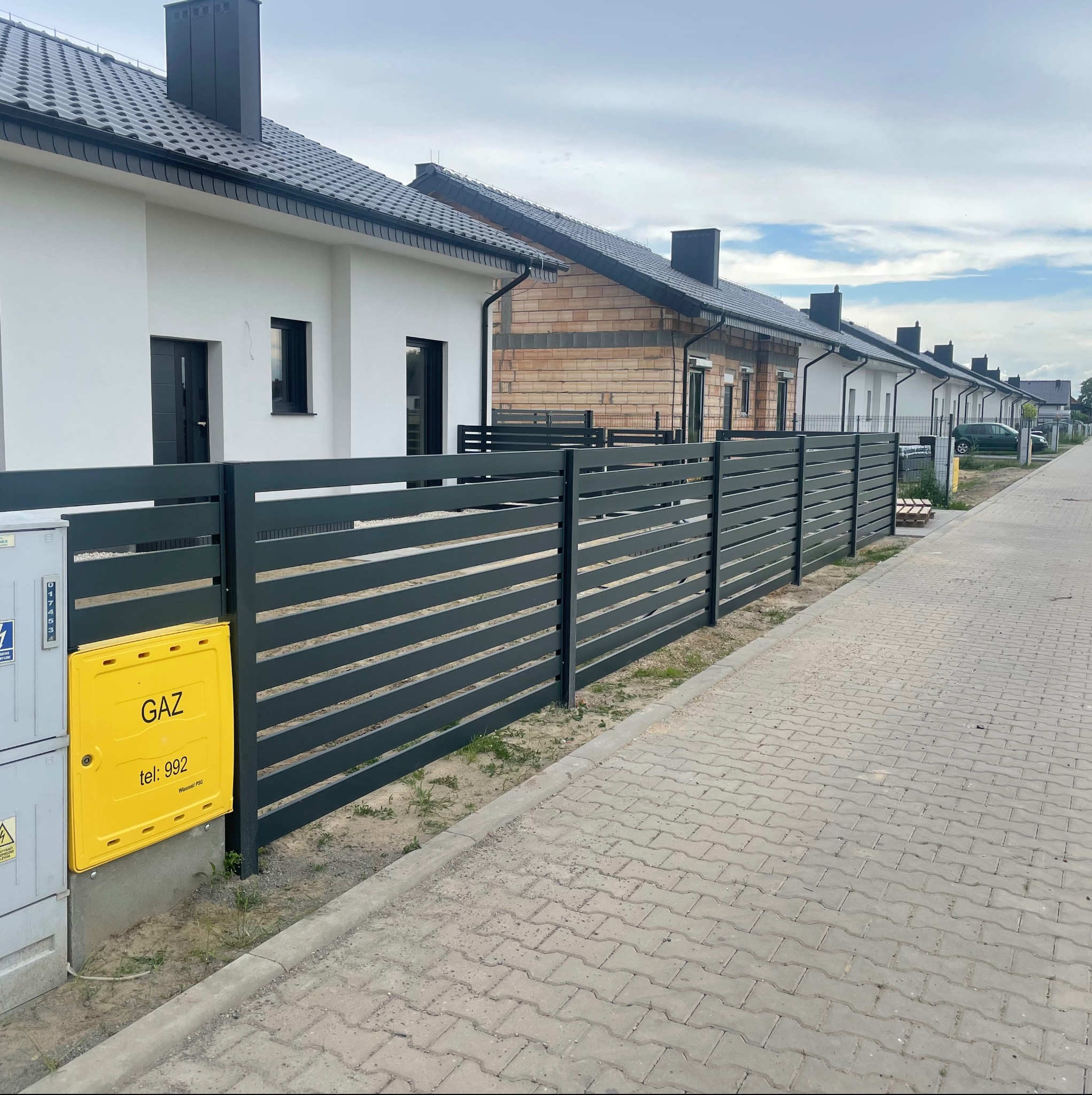 Szary, poziomy płot panelowy na tle szeregu domów jednorodzinnych w zabudowie szeregowej. Widoczna skrzynka gazowa. Ujęcie z poziomu chodnika.