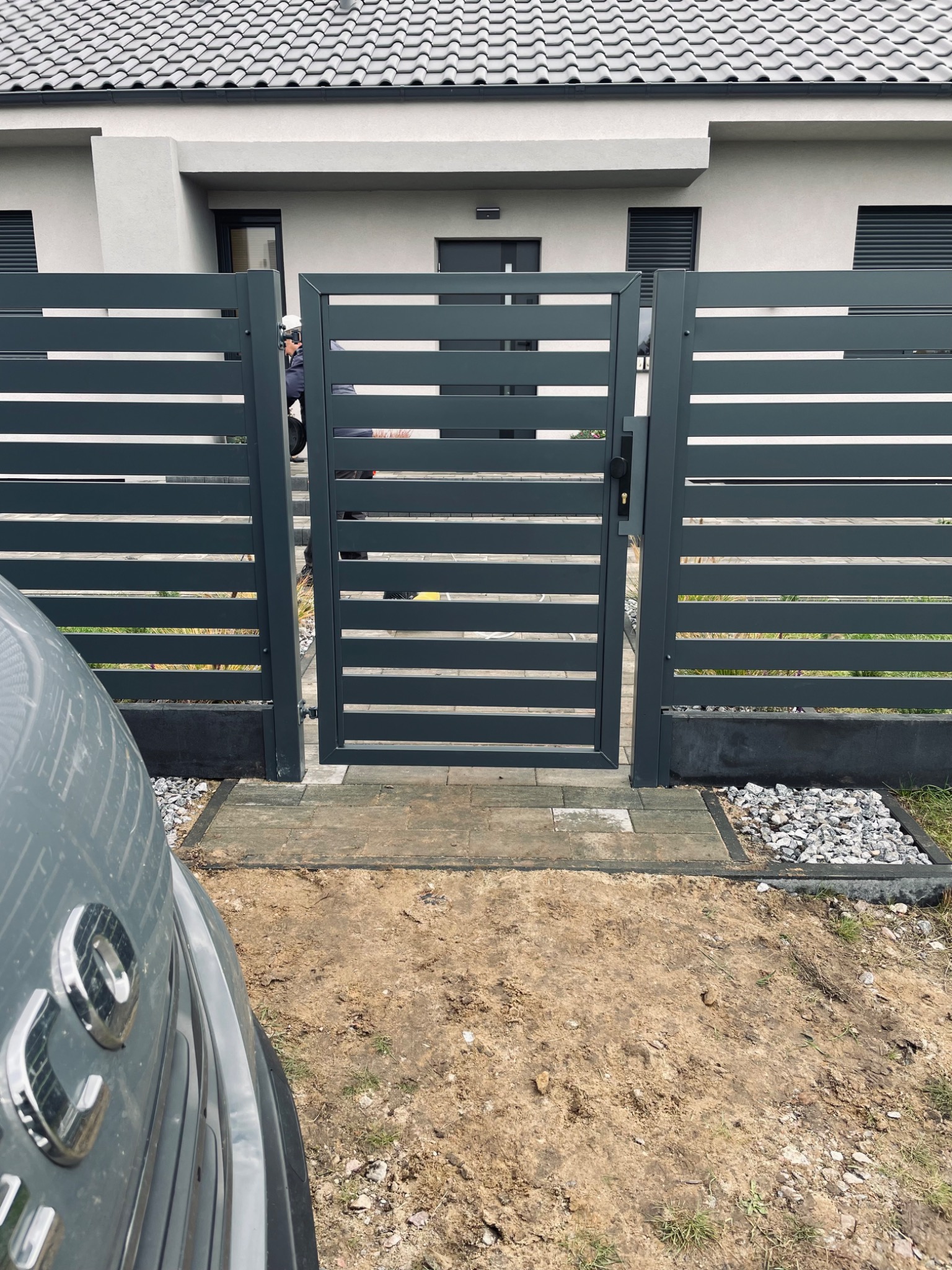 Nowoczesna, pozioma brama metalowa w kolorze antracytowym, z widocznym fragmentem samochodu po lewej stronie. Tło stanowi elewacja domu z szarą dachówką.