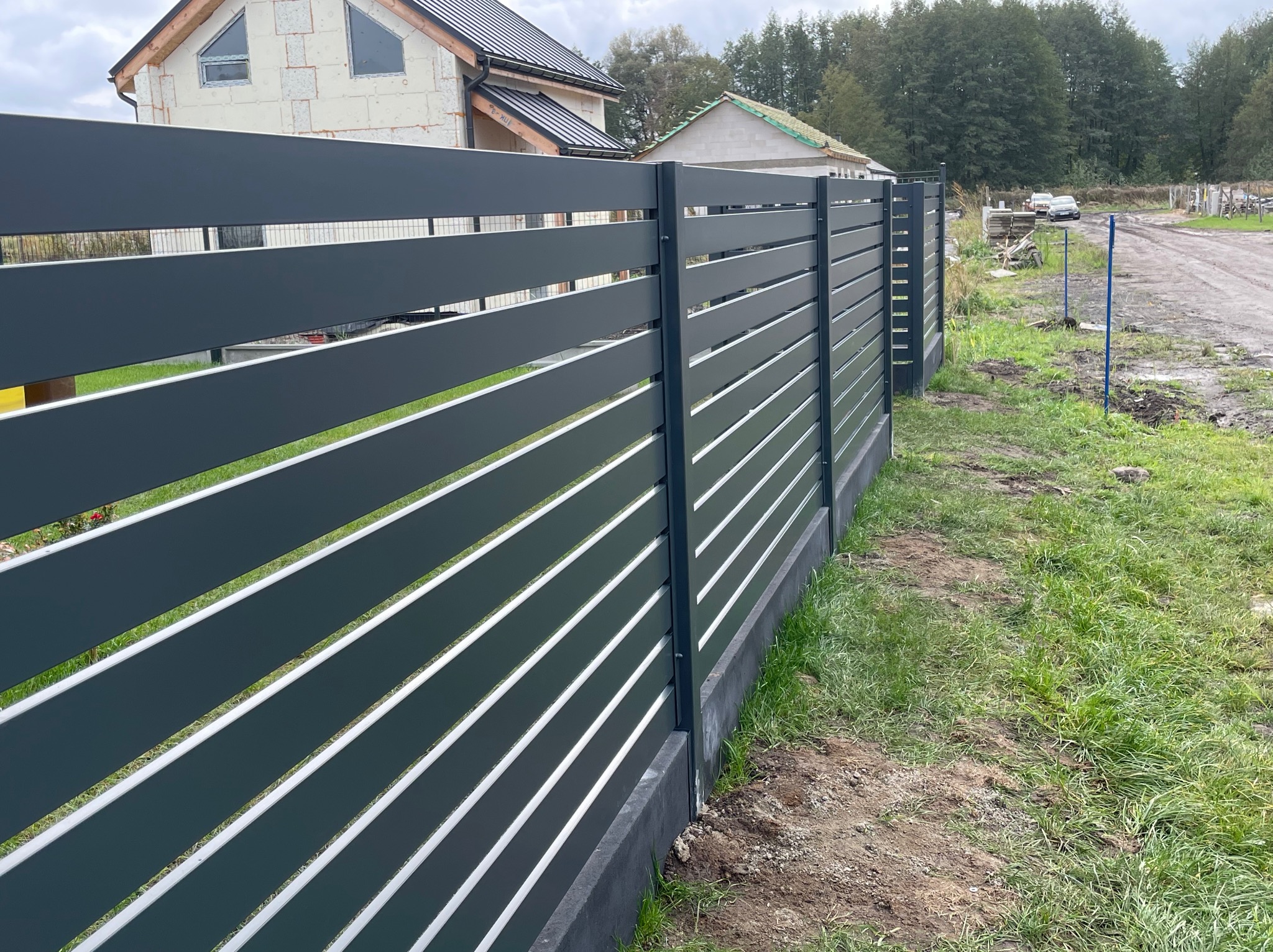 Nowoczesne, poziome ogrodzenie metalowe w kolorze grafitowym, z widocznymi prześwitami, na tle trawnika i budynków w trakcie budowy. Ujęcie z niskiej perspektywy.