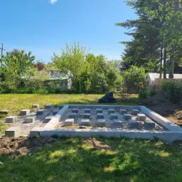 Betonowa rama fundamentowa z równo rozmieszczonymi betonowymi bloczkami, przygotowana pod budowę małego domku na zielonej działce w słoneczny dzień.