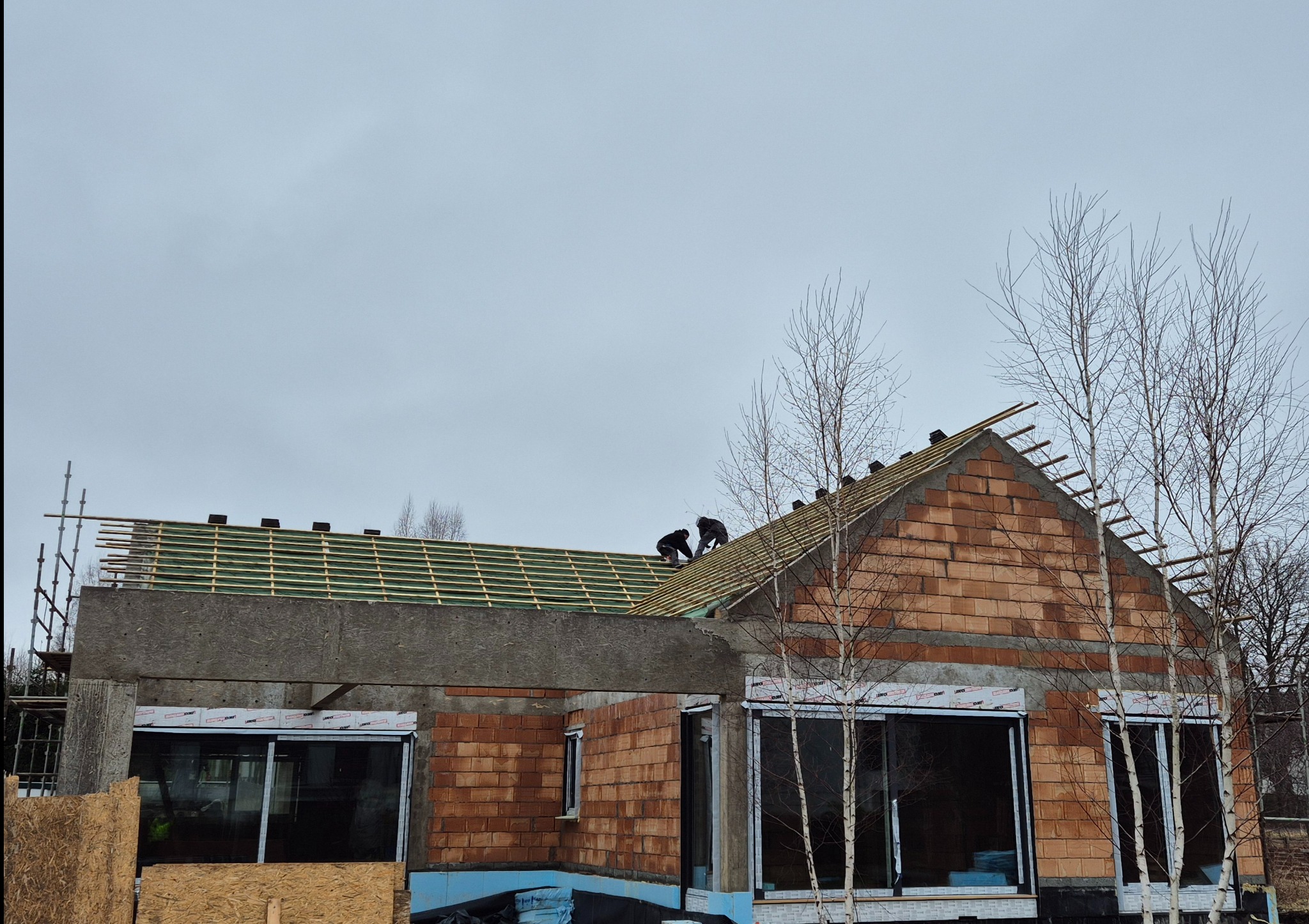 Budowa domu z czerwonej cegły w Koszalinie. Widoczni dwaj robotnicy na dachu, montujący konstrukcję. Stan surowy otwarty. W tle pochmurne niebo.