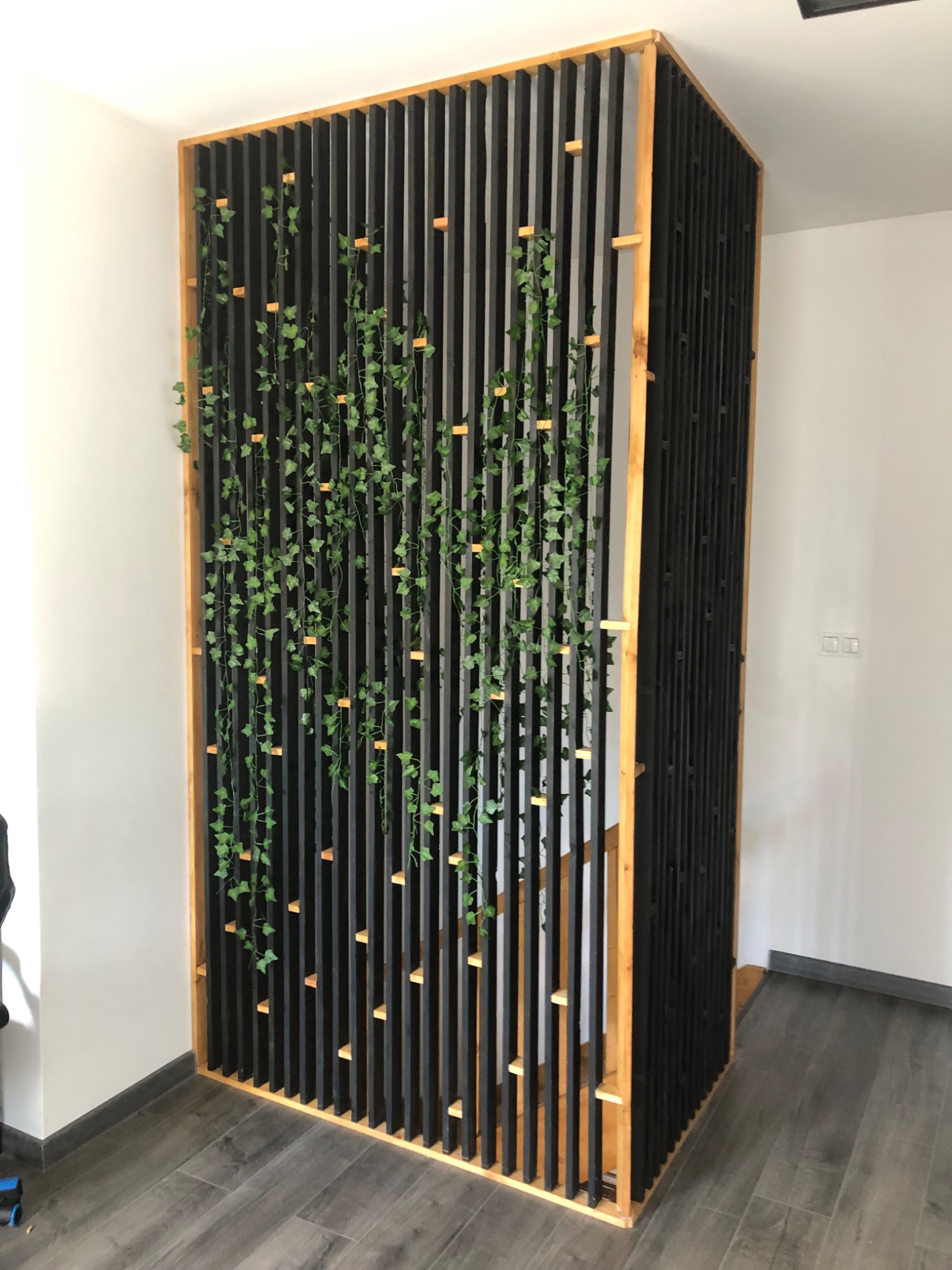 Drewniana ażurowa ścianka działowa w czarnym kolorze z elementami naturalnego drewna, ozdobiona pnącą zielenią, oddzielająca schody od reszty pomieszczenia.