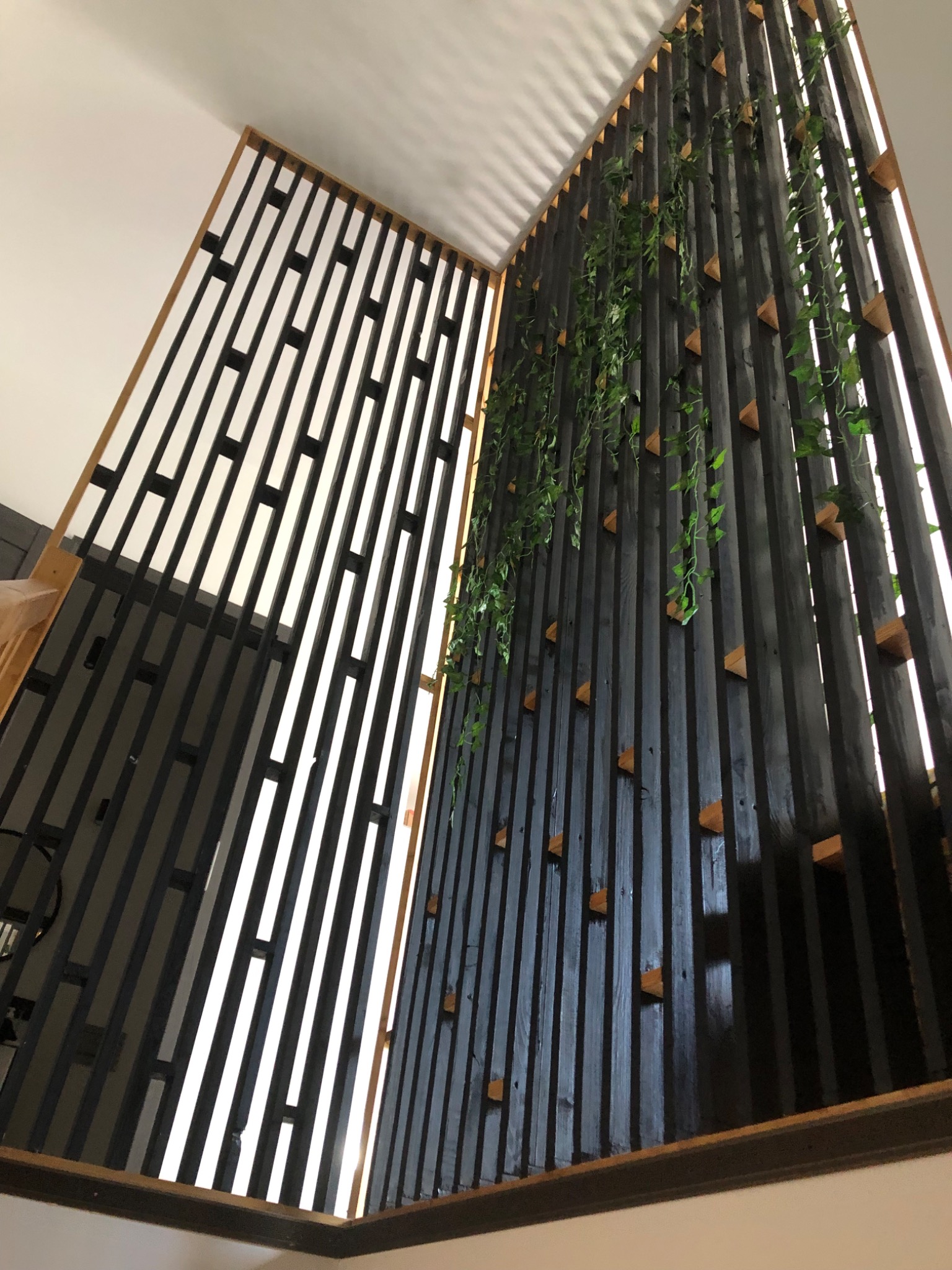 Wysoka, ażurowa ścianka działowa z pionowych, czarnych listew z dekoracyjnymi, drewnianymi elementami w kształcie trójkątów i zwisającą sztuczną roślinnością, widok z dołu.