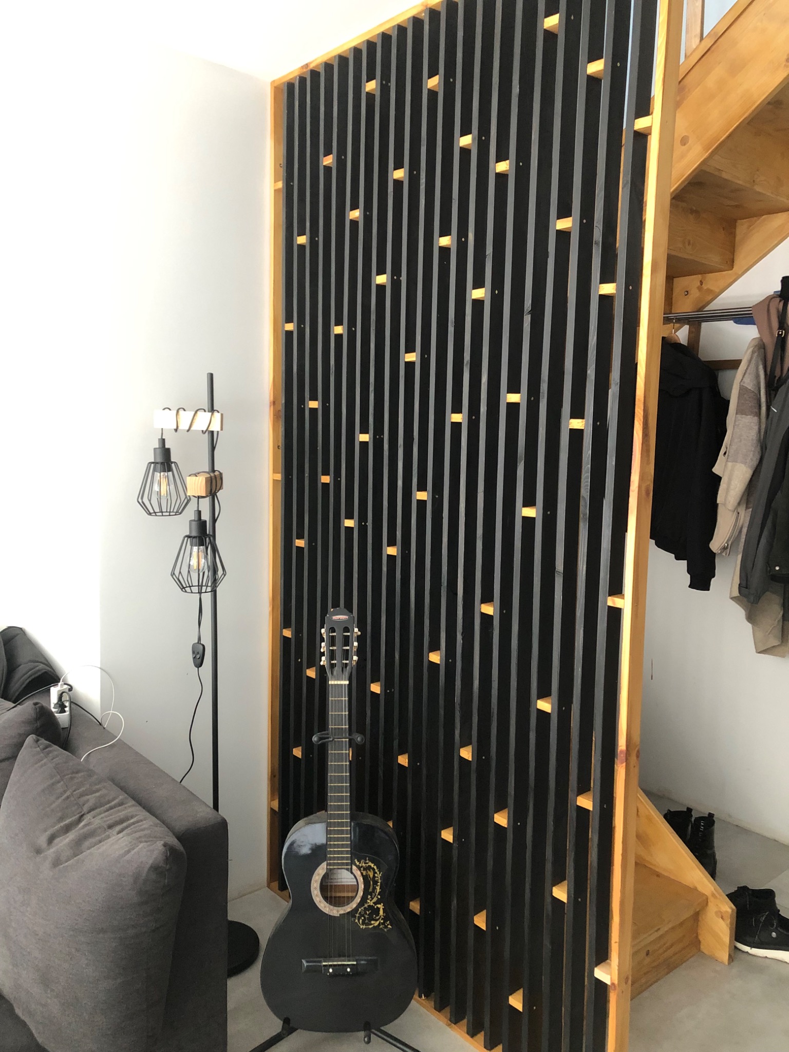 Nowoczesna, ażurowa ścianka działowa z czarnych listew drewnianych na jasnym drewnianym stelażu, gitara akustyczna oparta o ściankę, obok szara sofa i lampa podłogowa, w tle schody.
