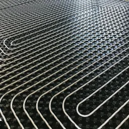 Białe rurki ułożone w regularnych odstępach na czarnej macie z wypustkami, instalacja w trakcie montażu, widoczne okno i fragment ściany.