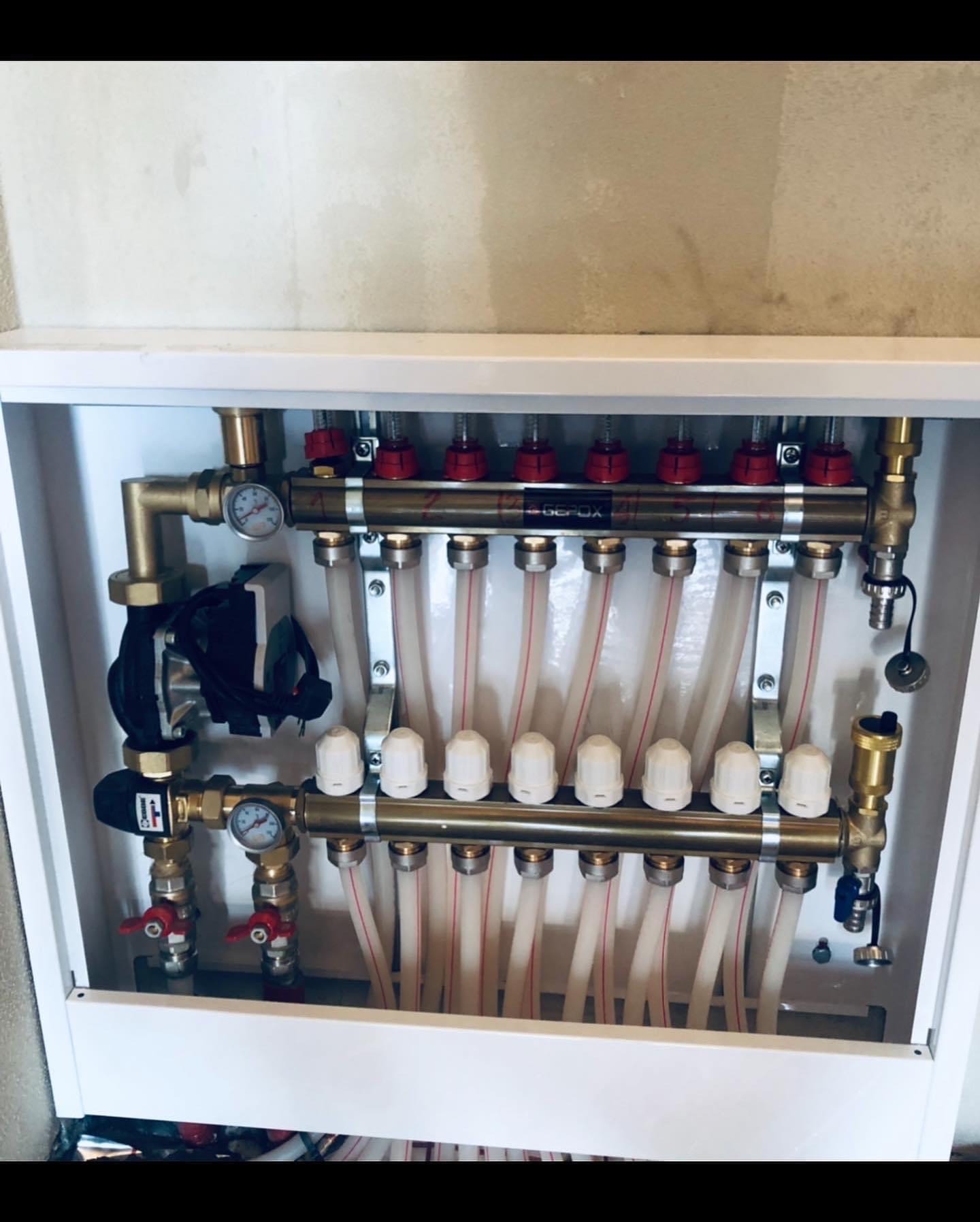 Rozdzielacz z pompą obiegową i rurami w szafce, instalacja w trakcie podłączania, widoczne manometry i zawory.
