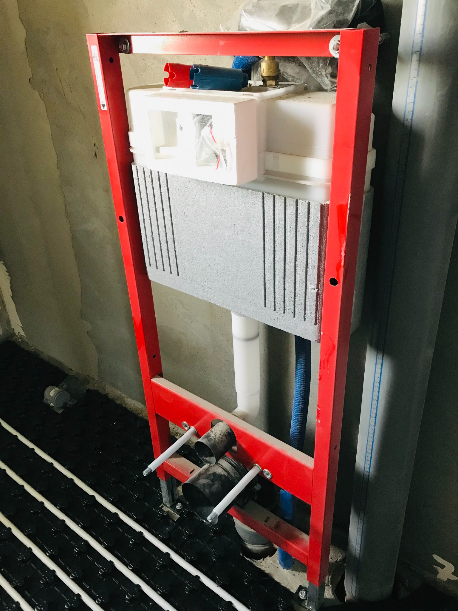 Stelaż podtynkowy WC w czerwonej ramie, zamontowany na tle surowej ściany, z widocznym fragmentem instalacji ogrzewania podłogowego w czarnej macie.