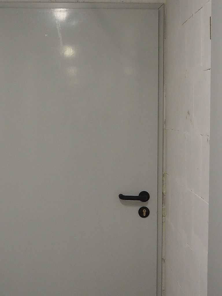Szare, gładkie skrzydło drzwiowe z czarną klamką i zamkiem, widoczne na tle pomalowanej ściany z drobnymi uszkodzeniami.