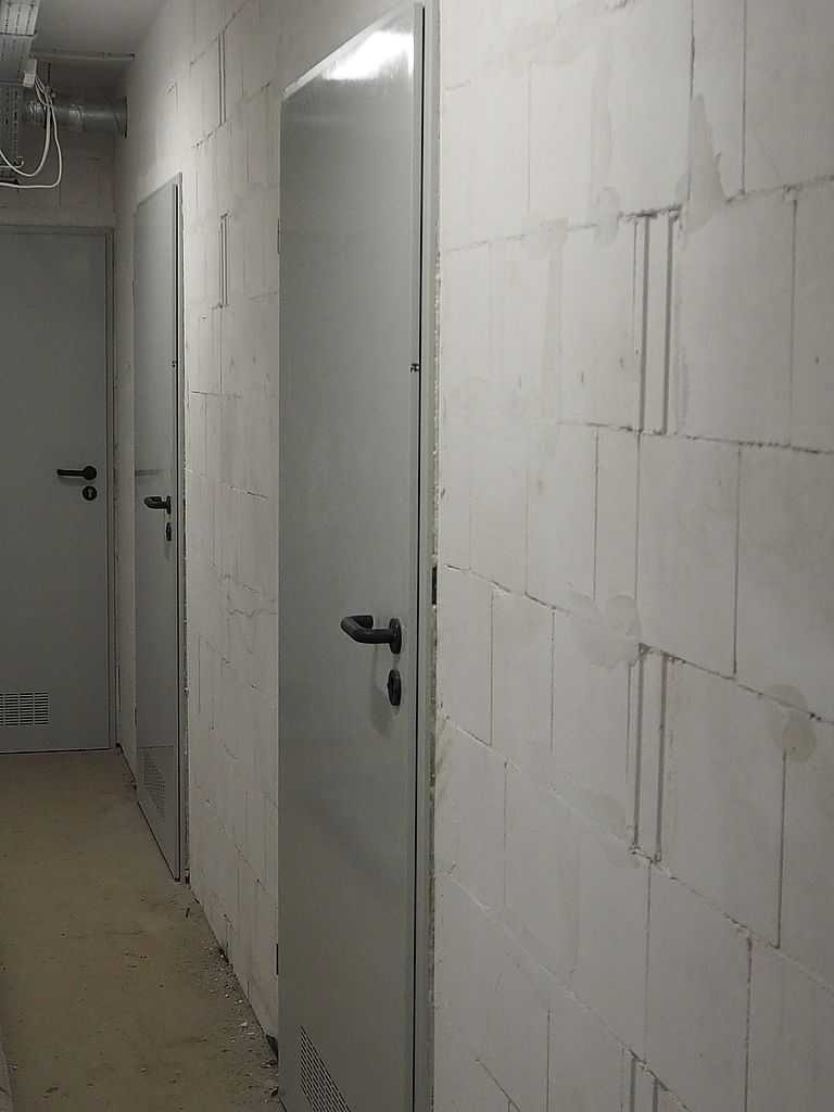 Szare drzwi techniczne Wiśniowski zamontowane w surowym wnętrzu z białymi ścianami z betonu komórkowego, widoczne przewody pod sufitem.