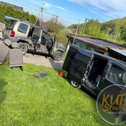 Dwa samochody na trawie, z wyjętymi fotelami samochodowymi obok; jeden samochód osobowy z otwartymi drzwiami i bagażnikiem, drugi to czarny van z otwartymi drzwiami, widoczne logo firmy nałożone...