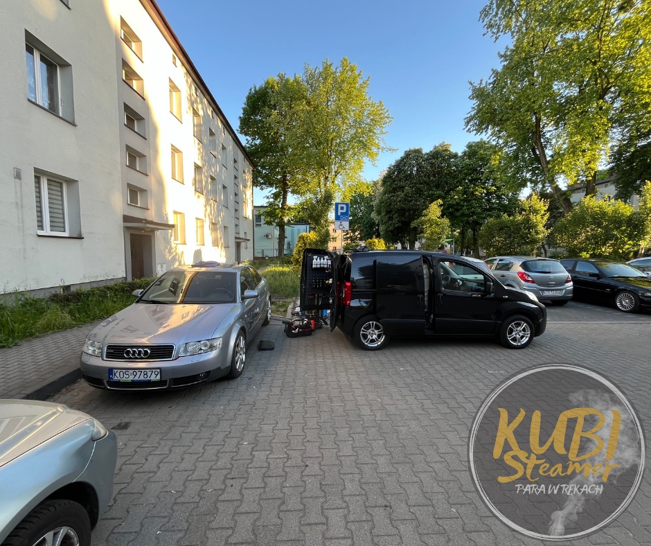 Czarna furgonetka z otwartymi drzwiami i rozłożonym sprzętem obok srebrnego Audi na parkingu; logo firmy w prawym dolnym rogu.