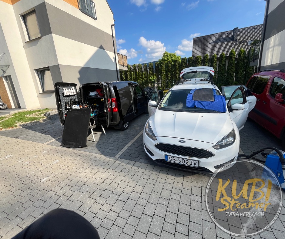 Mobilna jednostka czyszcząca z otwartymi drzwiami, wyposażona w sprzęt do czyszczenia, obok białego samochodu z otwartymi drzwiami, na którego masce leży niebieska ściereczka i dywanik. W tle...