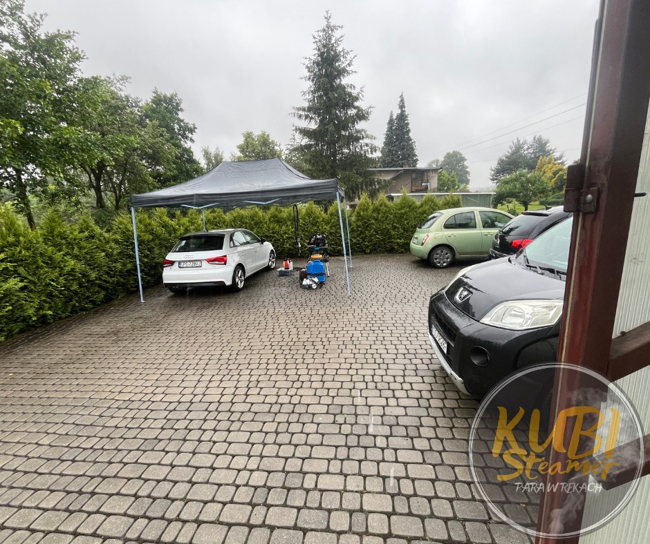 Na zewnątrz, na brukowanym podjeździe, białe Audi A3 pod szarym namiotem typu pop-up, obok stoi sprzęt do czyszczenia parowego; w tle żywopłot i inne zaparkowane samochody. W rogu widoczne logo firmy.