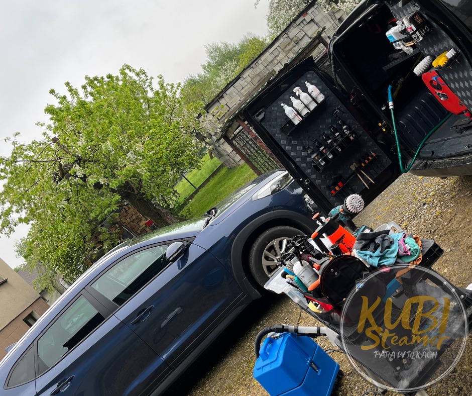 Mobilna myjnia parowa: Niebieski SUV, otwarte drzwi bagażnika z uporządkowanym zestawem środków czyszczących i narzędzi, wózek z akcesoriami do czyszczenia tapicerki, logo firmy Kubi Steamer...