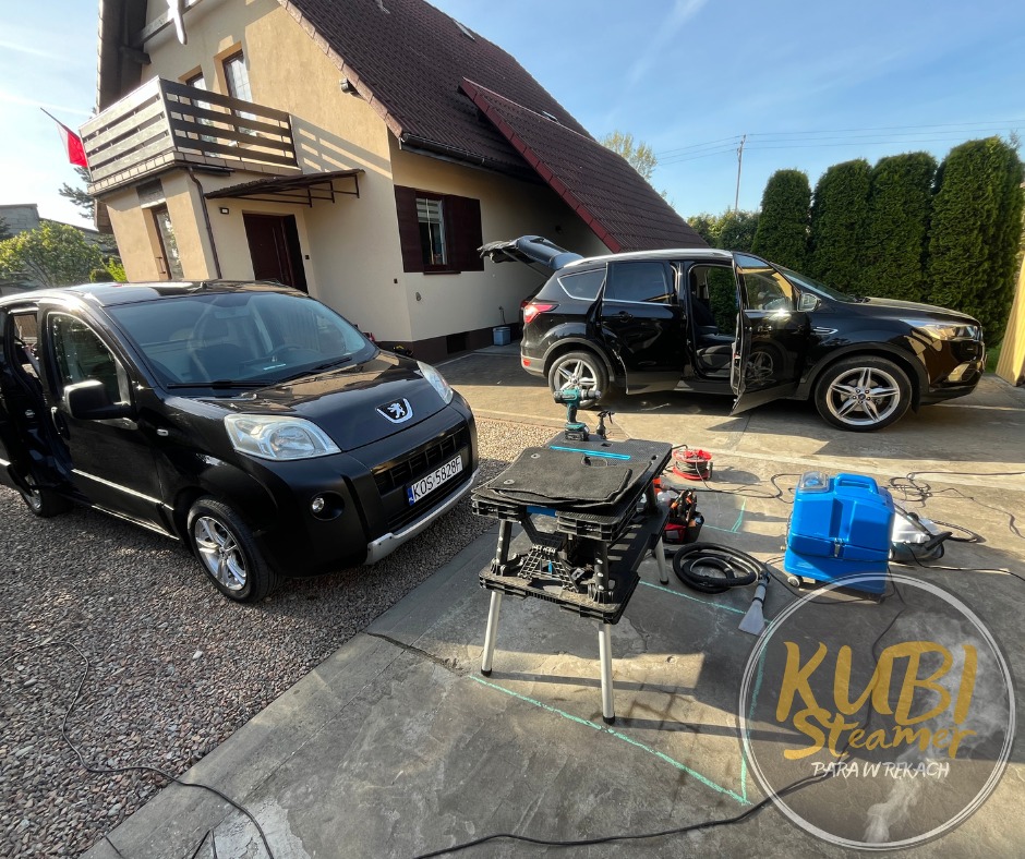 Dwa czarne samochody z otwartymi drzwiami przed domem; widoczny sprzęt do czyszczenia, w tym niebieski odkurzacz piorący i stolik z dywanikami samochodowymi.