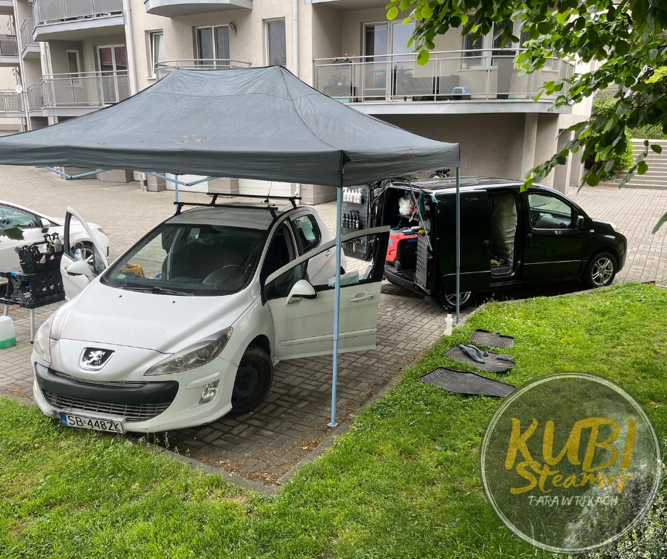 Mobilna usługa czyszczenia wnętrza samochodu na zewnątrz budynku: biały Peugeot 308 z otwartymi drzwiami pod szarym namiotem, czarny van z otwartymi drzwiami prezentujący sprzęt do czyszczenia...