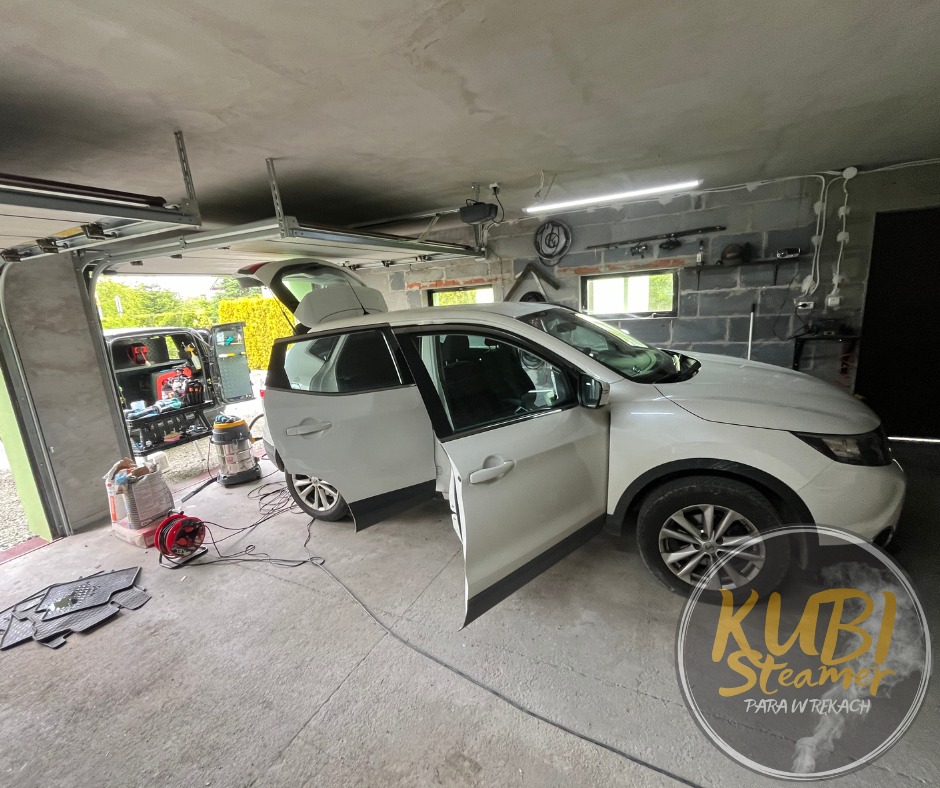 Biały samochód z otwartymi drzwiami i bagażnikiem w garażu, widoczny sprzęt do czyszczenia, w tle otwarte drzwi garażowe z zielenią na zewnątrz, w prawym dolnym rogu logo firmy.