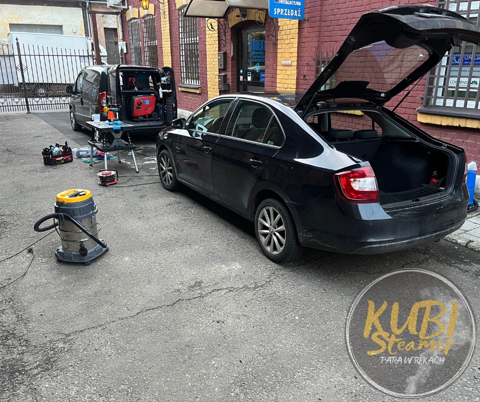 Czarny samochód z otwartym bagażnikiem, obok zaparkowany van z wyposażeniem do czyszczenia, w tle budynek z cegły, widoczny odkurzacz przemysłowy i narzędzia do czyszczenia, usługa mobilna.