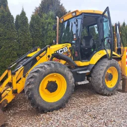 Żółta koparko-ładowarka JCB zaparkowana na żwirowym podłożu, z otwartymi drzwiami kabiny i widocznymi elementami hydraulicznymi, na tle wysokich, zielonych tuj.