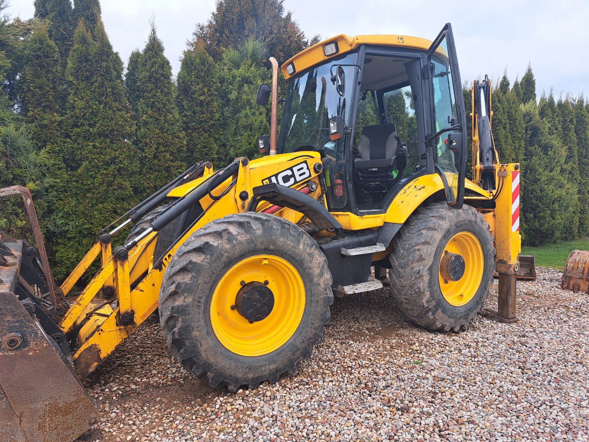 Żółta koparko-ładowarka JCB zaparkowana na żwirowym podłożu, z otwartymi drzwiami kabiny i widocznymi elementami hydraulicznymi, na tle wysokich, zielonych tuj.